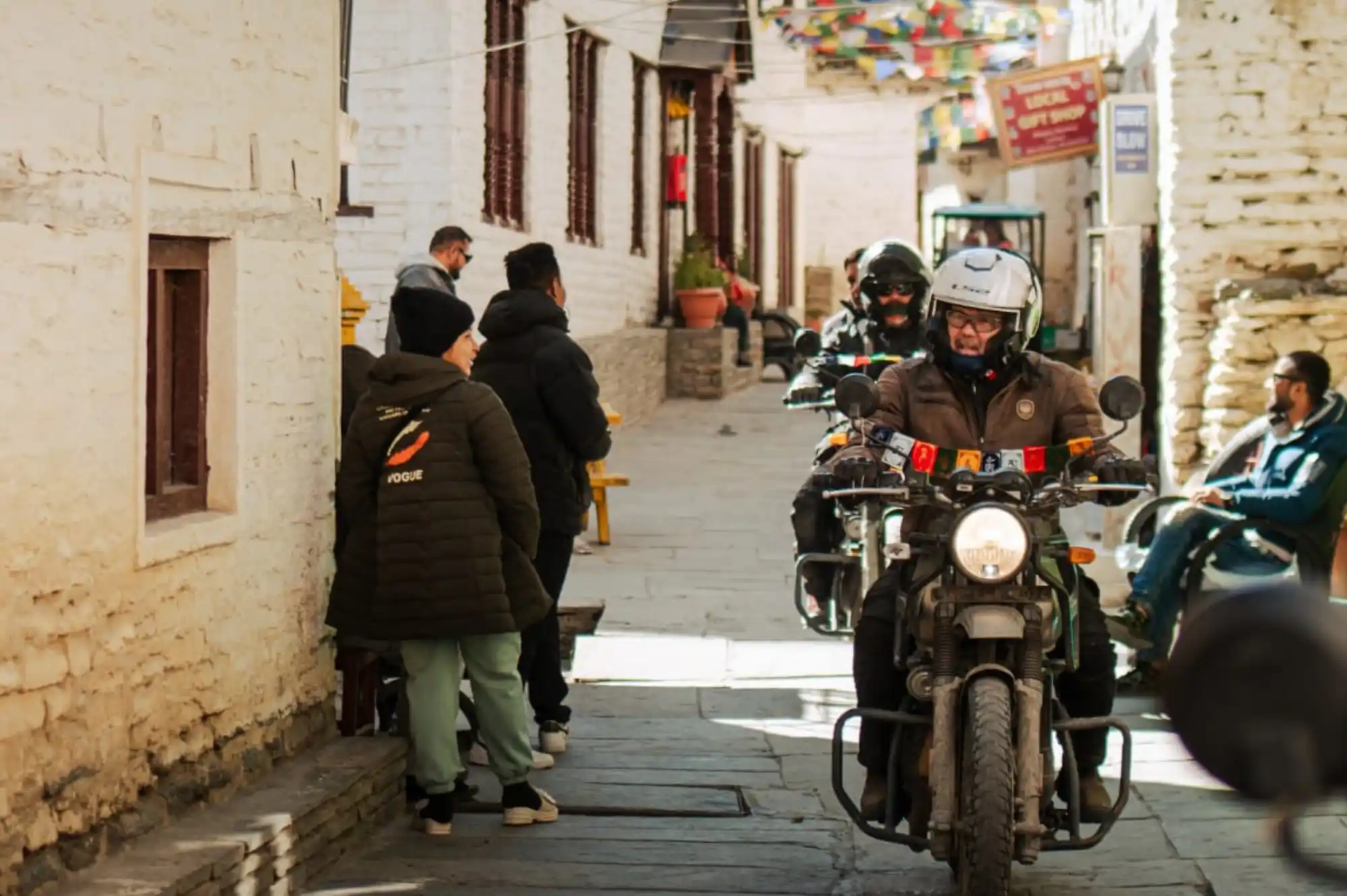 Motorbike riders in full gear navigate a narrow, traditional cobblestone street in a village, with white stone buildings and prayer flags overhead, typical of Mustang region on a motorbike tour.