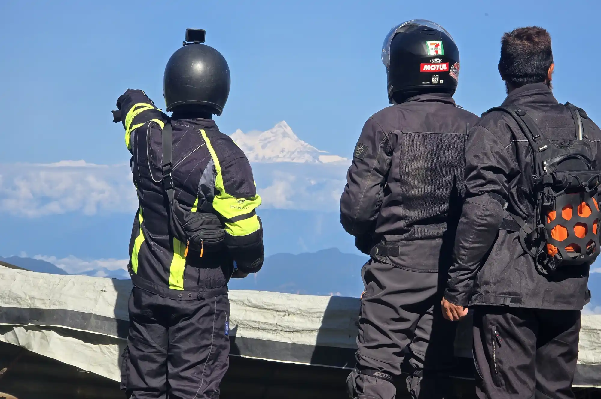 Three motorbike tourists in full riding gear stand with their backs to the camera, looking out at a magnificent snow-capped mountain peak partially obscured by clouds, showcasing a rewarding view during a motorbike tour.