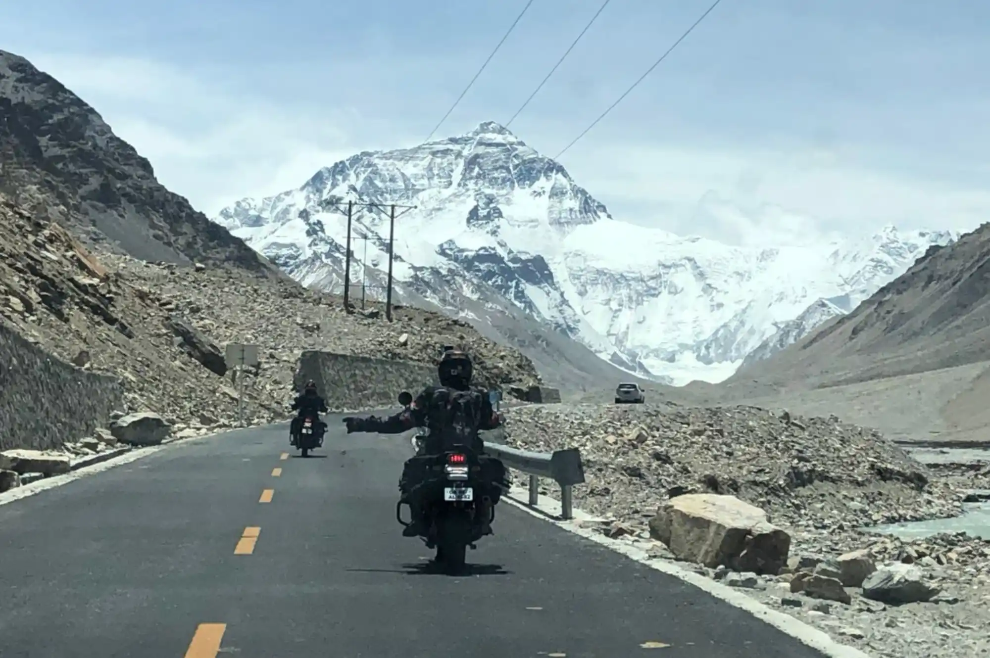 A motorcyclist riding on a paved road with arms outstretched, facing away from the camera, with the majestic snow-capped peak of Mount Everest visible in the distance under a blue sky, an iconic view on motorbike tours to Everest Base Camp.