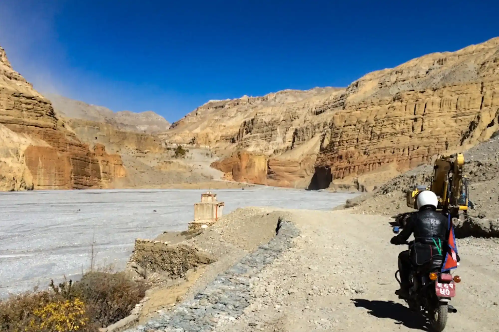 A motorcyclist rides on a dirt track beside a wide, dry riverbed, with towering, eroded canyon walls under a clear blue sky, capturing the adventurous spirit of motorbike tours in Upper Mustang.