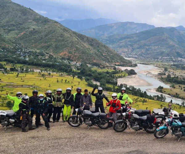 A happy group of riders poses with their motorcycles on a viewpoint overlooking a picturesque Nepalese valley with a winding river and terraced fields.