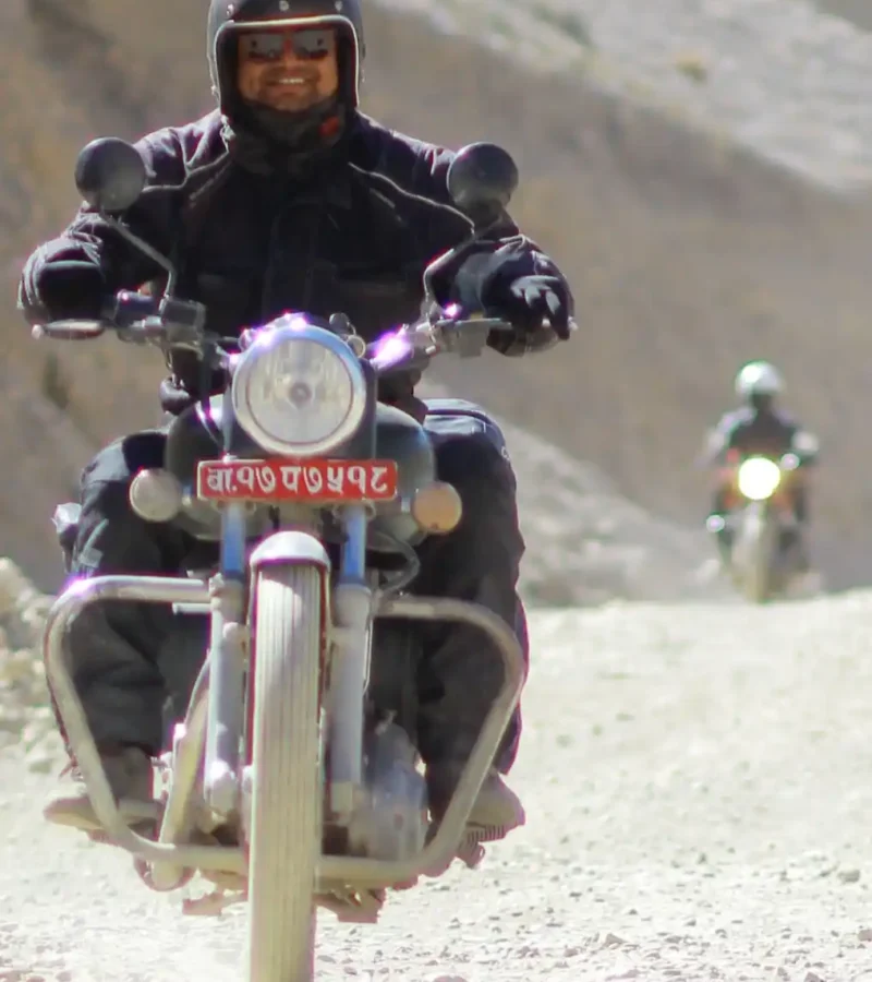A smiling motorcyclist in full gear, wearing a helmet with an action camera, rides a Royal Enfield motorbike directly towards the camera on a dusty, unpaved road in a barren, mountainous landscape, with another rider in the distance, characteristic of motorbike tours in Upper Mustang, Nepal.