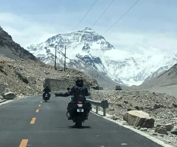 A motorcyclist riding on a paved road with arms outstretched, facing away from the camera, with the majestic snow-capped peak of Mount Everest visible in the distance under a blue sky, an iconic view on motorbike tours to Everest Base Camp.